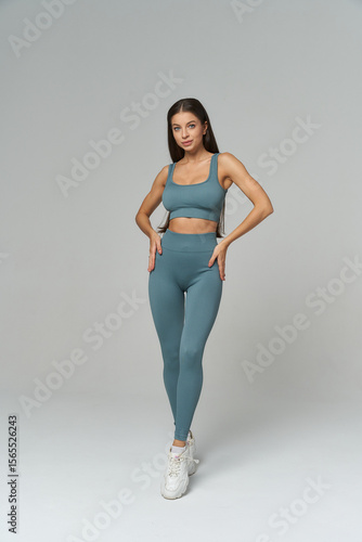 Fitness model in teal activewear, standing with a confident pose. She wears a sports bra and leggings, paired with white sneakers. Her long dark hair is sleek and straight, hands on hips