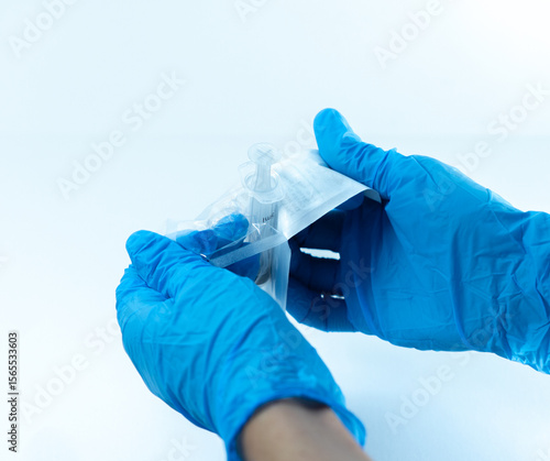 Wide-angle view of gloved hands opening a disposable syringe from its sterile blister packaging. Sterile medical preparation in clinical or hospital setting.