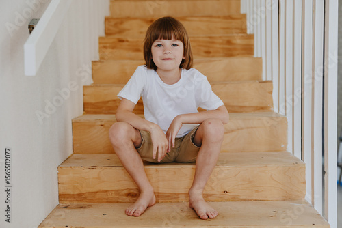 Fotografie Happy boy sitting on wooden stairs, wearing a white t-shirt and shorts