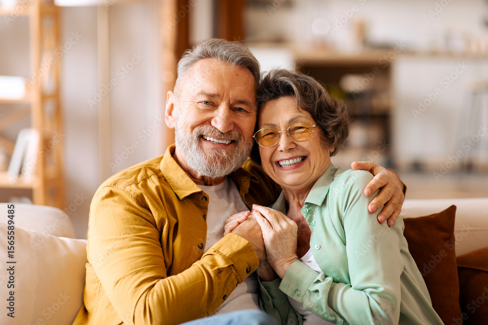 Obraz premium Happy elderly partners. Senior man and woman sharing tender moment, hugging and radiating happiness, sitting on sofa in living room and smiling at camera