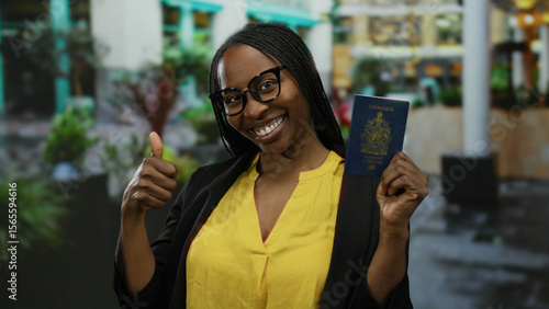 Woman outdoors in city holding canadian passport smiling with thumbs up gesture wearing glasses and yellow shirt against urban backdrop conveying travel approval.
