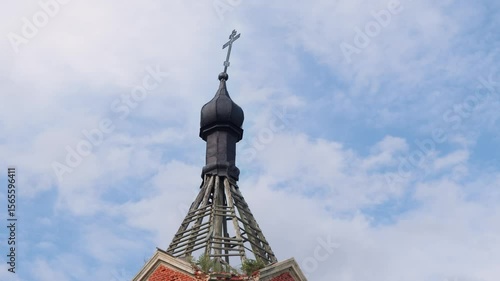 A close-up of a small black Orthodox dome with a lopsided cross on top of a brick-and-wooden abandoned bell tower against background of blue sky with clouds.