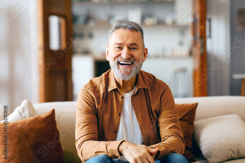 Portrait of Excited senior man posing alone at home, sitting on couch and smiling, looking at camera in living room