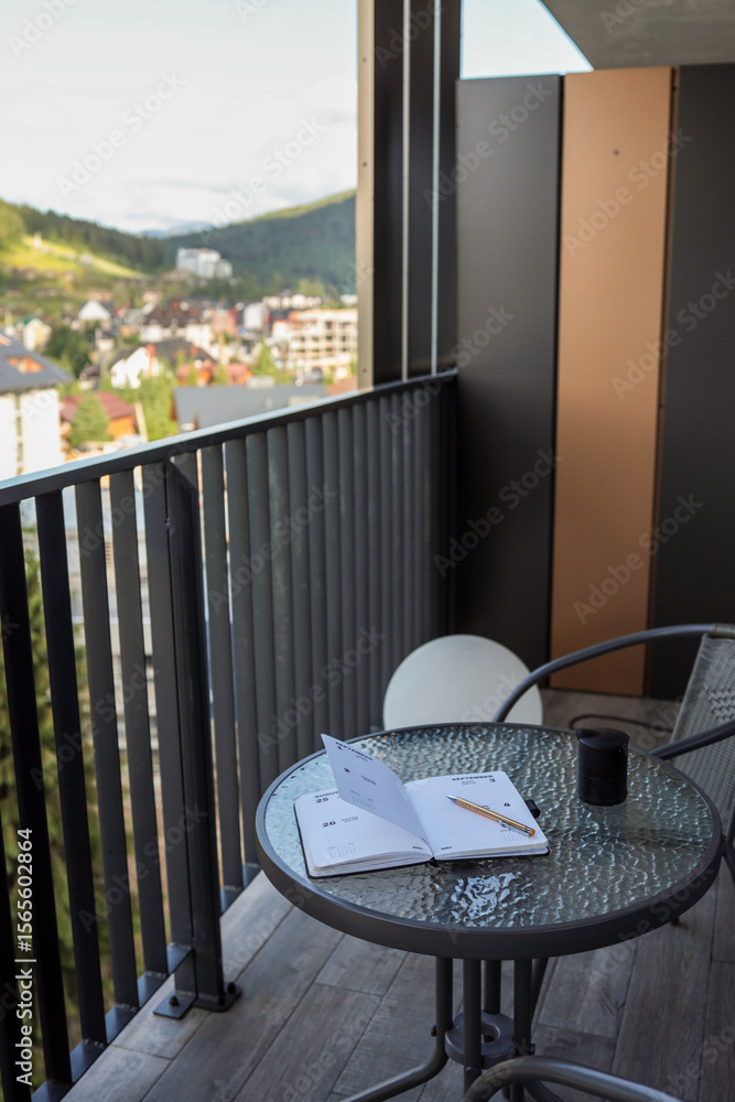 Obraz premium Glass table on balcony with open planner, pen, and coffee cup. Modern urban view with city buildings and mountains. Concept of planning, relaxation, remote work, productivity, lifestyle