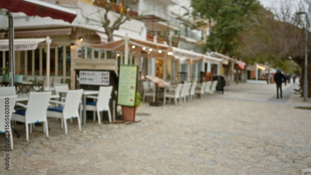 Obraz premium Defocused promenade in mallorca with blurred chairs and tables lining a mediterranean walk under soft lighting, depicting a peaceful and inviting outdoor dining scene.