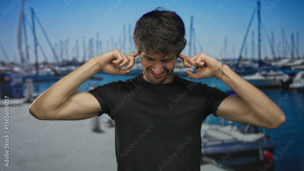 Fototapeta premium Man covers ears with hands at port pier by waterfront and moored boats under bright sky; annoyance.