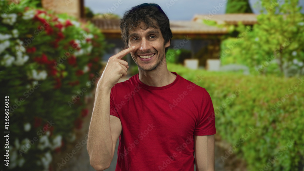 Obraz premium Young man wearing red shirt pointing finger to cheek in lush green park with hedges and blossoms under bright daylight; happiness.
