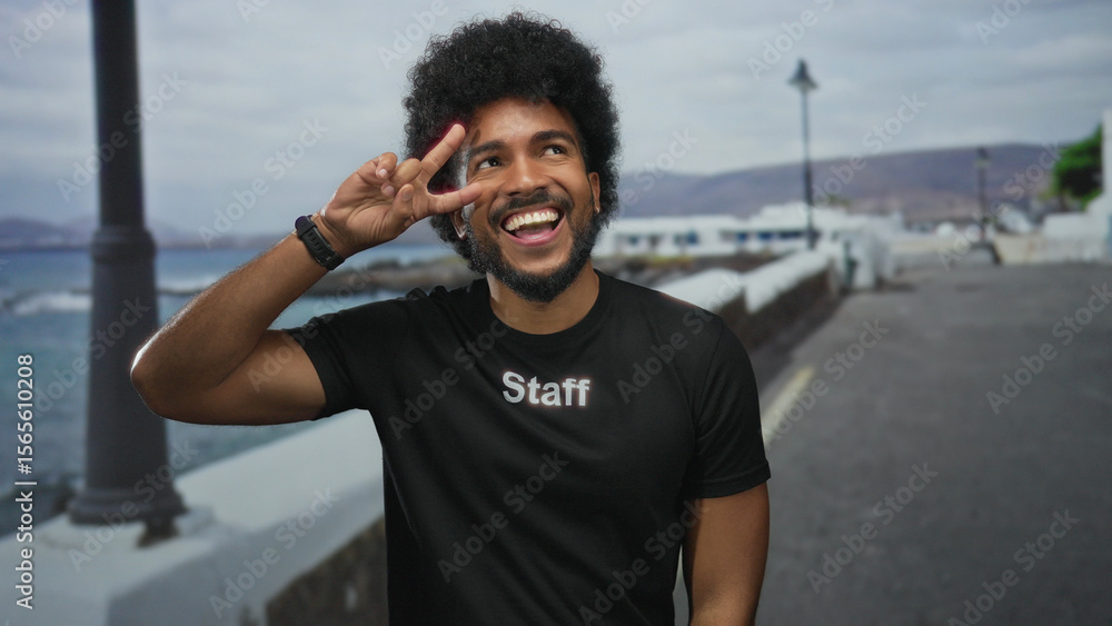 Naklejka premium African american man in staff uniform makes victory gesture outdoors on seaside promenade, smiling brightly under cloudy skies with serene sea view.
