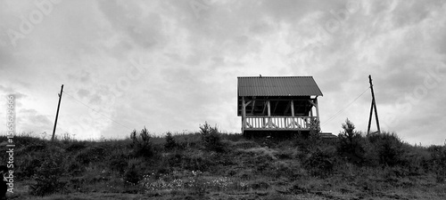Black and white photo of a solitary wooden cabin on a hill against a cloudy sky. The scene conveys solitude and tranquility, with sparse vegetation.