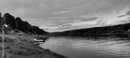 A serene black-and-white riverside scene with a small boat on grassy banks, calm water reflecting a cloudy sky, and trees lining the opposite shore.