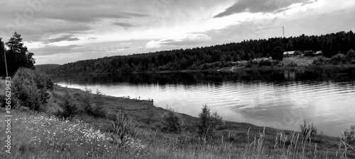 A serene black and white landscape shows a calm river reflecting the cloudy sky, bordered by grassy fields and dense trees, creating a tranquil atmosphere.