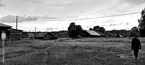 A black-and-white image shows a solitary figure walking through a grassy field. In the distance, old, rustic buildings under a cloudy sky evoke a somber mood.
