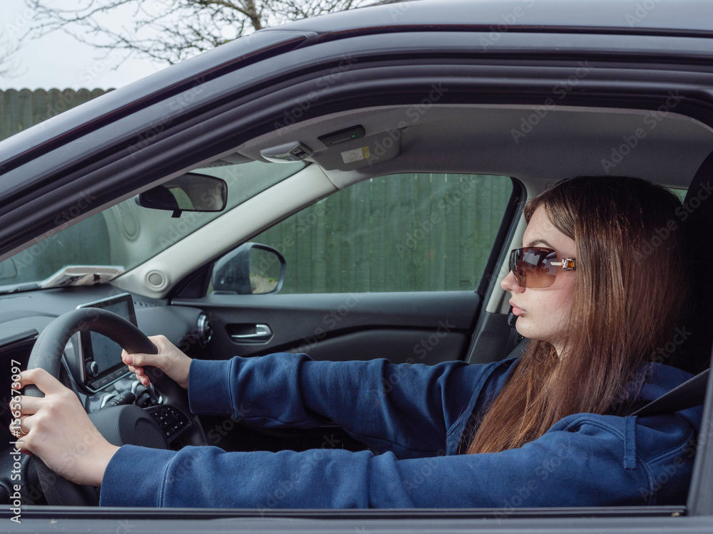 Naklejka premium A young inexperienced woman is driving a car with her sunglasses on. She is wearing a blue hoodie. High cost of insurance for young drivers concept. Travel and tourism concept.