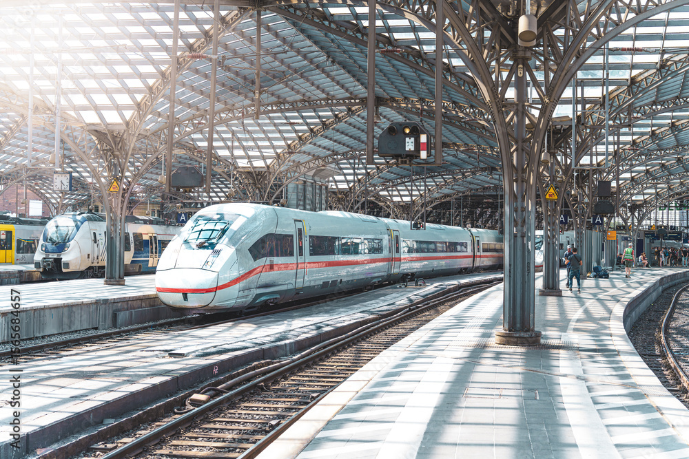 Fototapeta premium On a sunny day, a train departs from Cologne Central Station. Alongside the rails, the pedestrian walkway, industrial architecture and a glass roof can be seen.