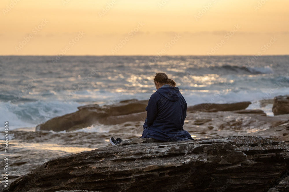 Silhouetted woman sits on rock at beach during sunrise moment Silhouetted woman sits on rock at beach during sunrise moment