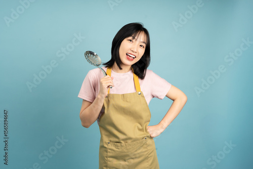 Young Asian woman in an apron holding a cooking utensil with a cheerful expression in a studio setting with a blue backdrop