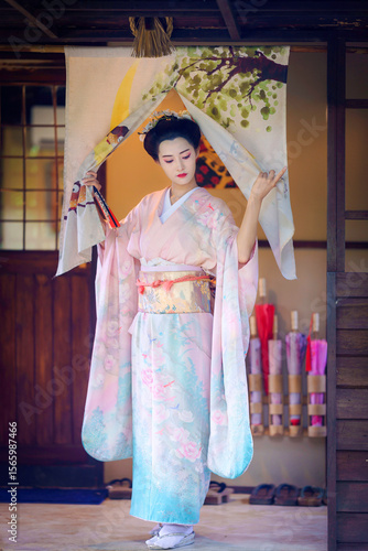 Pretty young Asian Japanese woman in a Traditional Furisode Kimono Yukata dress costume. A young Asian traveller girl stands and parts the white curtain in front of the house door.