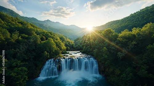 Lush green forest cascades down to a vibrant turquoise waterfall under a bright, sunny sky, creating a serene and picturesque natural scene. The light filters through the trees, highlighting the