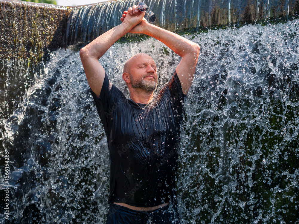 Fototapeta premium A bald man is standing under a waterfall, getting wet. Concept of relaxation and rejuvenation, as the man is enjoying the refreshing water. Model is slim and in good physical form. Male in his 50s