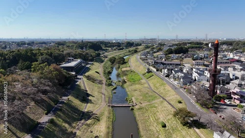 Rising aerial drone shot of a serene riverside park in Japan, showing a small river flowing between manicured green embankments, walking paths, a distinctive orange clock tower, residential houses, an
