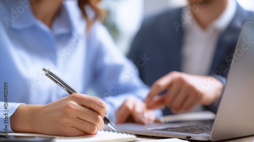 Close-up on a woman in a blue shirt writing in a notebook while a man in a suit points at her laptop. Focus on their hands and the work. 