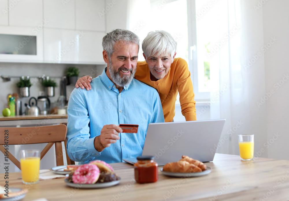 Portrait of an elderly senior couple having breakfast and looking at a laptop using a credit card for online shopping at home. Happy healthy affectionate senior couple eating and sitting at kitchen ta Portrait of an elderly senior couple having breakfast and looking at a laptop using a credit card for online shopping at home. Happy healthy affectionate senior couple eating and sitting at kitchen ta