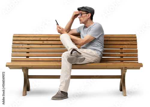 Mature man in his 50s sitting on a wooden bench and using a smartphone, studio shot isolated on white background. Casual clothing, wearing reading glasses and a hat