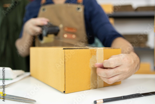A man is packing things into boxes. He uses scotch tape and string to tie the boxes, writes information on the boxes, and uses his mobile phone to take photos to record the product information.
