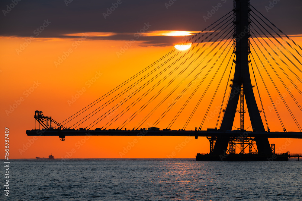 Fototapeta premium Fiery sunset casting an orange glow over the under-construction Danshui cable bridge in New Taipei Taiwan, creating a dramatic silhouette.