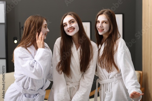 Happy women with facial masks in bathrobes posing after spa procedures indoors