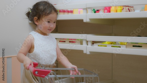 A young child explores a toy kitchen, engaging with colorful play food and a shopping cart in a bright playroom.