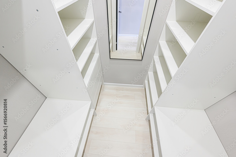 Fototapeta premium bright, clean walk-in closet featuring white shelving units on either side of a light wood floor, with a full-length mirror at the end of the hallway