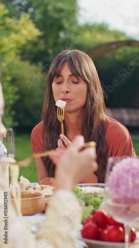 Charming Caucasian woman in beautiful dress sitting at outdoor table. Picking food with fork. Enjoying dinner near flowers and wine bottles. Stylish garden party. Vertical video orientation.