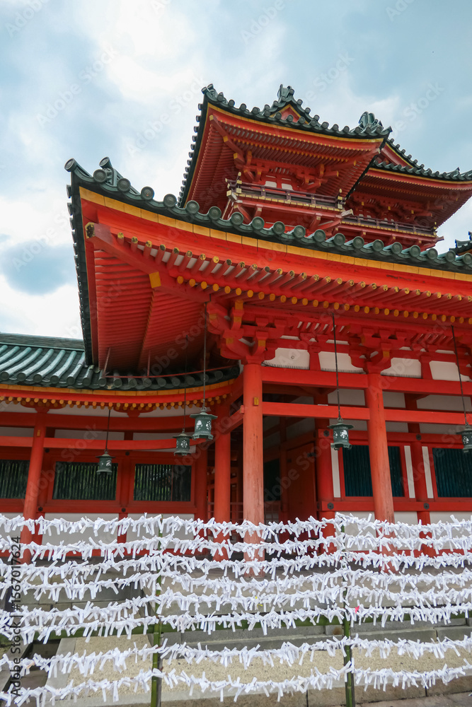 Naklejka premium Countless white paper fortunes, or omikuji, are tied in rows before a vibrant red shrine building, a popular spiritual tradition representing the hopes and prayers of visitors in Kyoto, Japan.