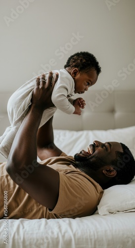 A father playfully lifts his baby son into the air while lying on a bed