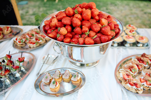 Outdoor strawberry buffet with silver bowls overflowing with ripe berries, surrounded by assorted mini desserts and savory snacks on a white tablecloth, creating a festive summer celebration ambiance