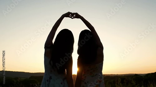 Two Young Women Create Heart Shape with Hands During Sunset in Field Silhouette