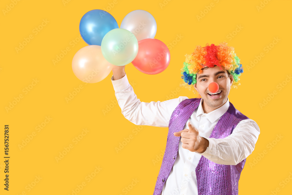 Fototapeta premium Happy young man in clown costume with balloons on yellow background