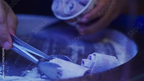 Close-up of syrup being drizzled over colorful rolled ice cream in a cup, served by vendor at vibrant night market.