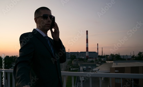 A silhouette of a man in a suit and tie, talking on the phone, against the background of a factory with pipes 