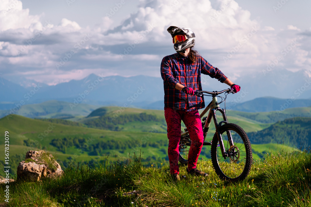 Obraz premium Athlete rider in bicycle helmet on his bike standing and resting before descent in mountains against the backdrop of epic mountains and landscape. MTB downhill discipline background.