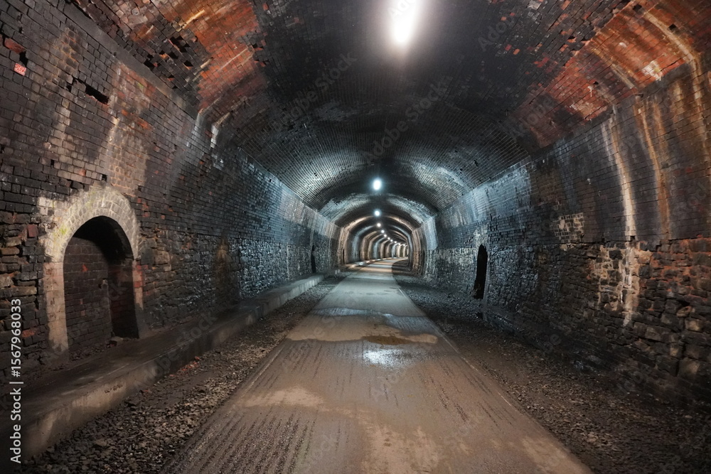 Fototapeta premium Historic brick railway tunnel with lights and wet gravel path in The Peak District, England.
