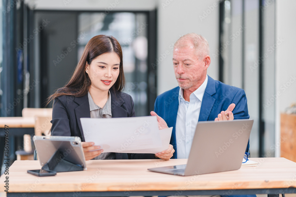 Young Asian businesswoman and senior Caucasian businessman working together in modern office, discussing documents and using laptop, teamwork and collaboration concept Young Asian businesswoman and senior Caucasian businessman working together in modern office, discussing documents and using laptop, teamwork and collaboration concept