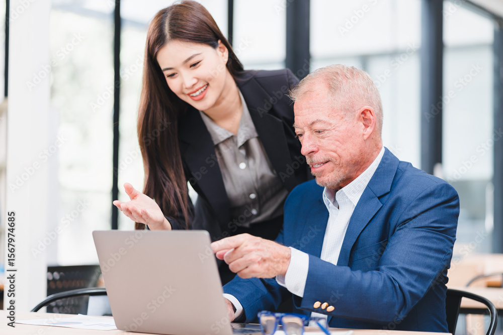 Obraz premium Young Asian businesswoman and senior Caucasian businessman working together in modern office, discussing documents and using laptop, teamwork and collaboration concept