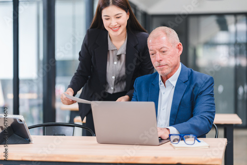 Young Asian businesswoman and senior Caucasian businessman working together in modern office, discussing documents and using laptop, teamwork and collaboration concept