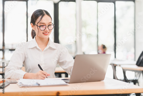 Young Asian woman, wearing glasses and white shirt, smiling while working on laptop and taking notes in modern bright office, professional and focused atmosphere