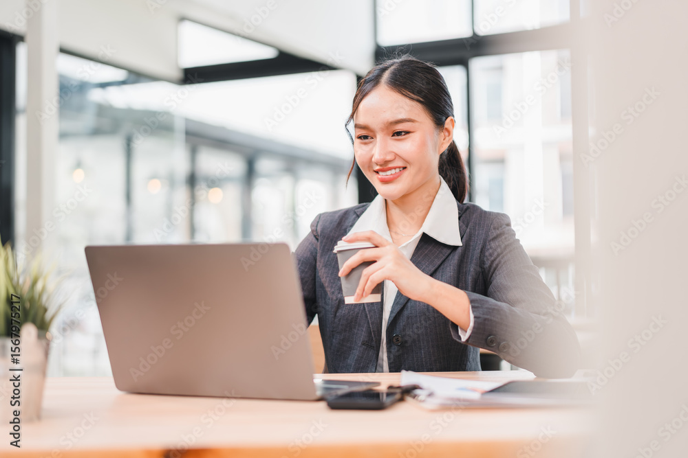 Fototapeta premium Young Asian businesswoman professional suit smiling working at modern office desk using laptop holding coffee cup natural daylight confident and productive atmosphere