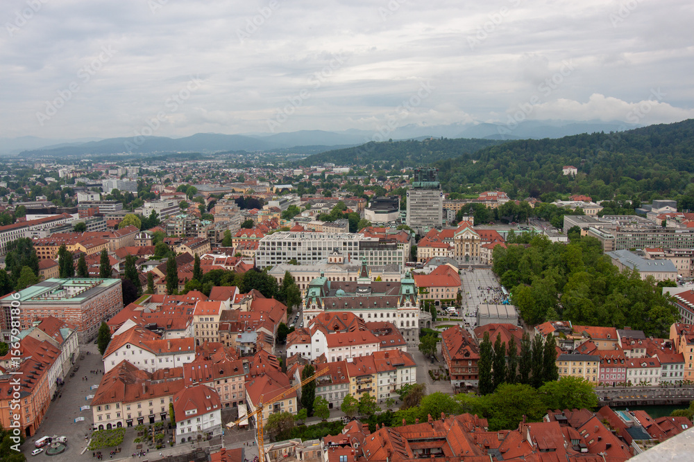 Fototapeta premium An expansive elevated view captures the cityscape of Ljubljana, on a cloudy day. Red-tiled rooftops dominate the foreground, showcasing the charming historic architecture of the old town
