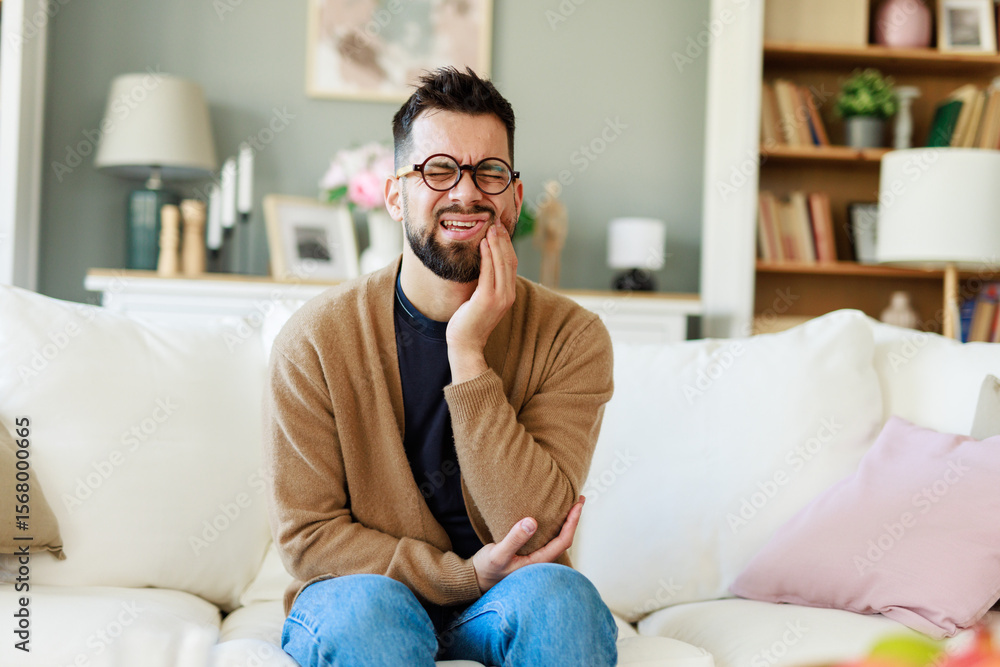 Young Man in Brown Cardigan Holding Jaw in Pain While Sitting on a Comfortable Living Room Sofa Young Man in Brown Cardigan Holding Jaw in Pain While Sitting on a Comfortable Living Room Sofa
