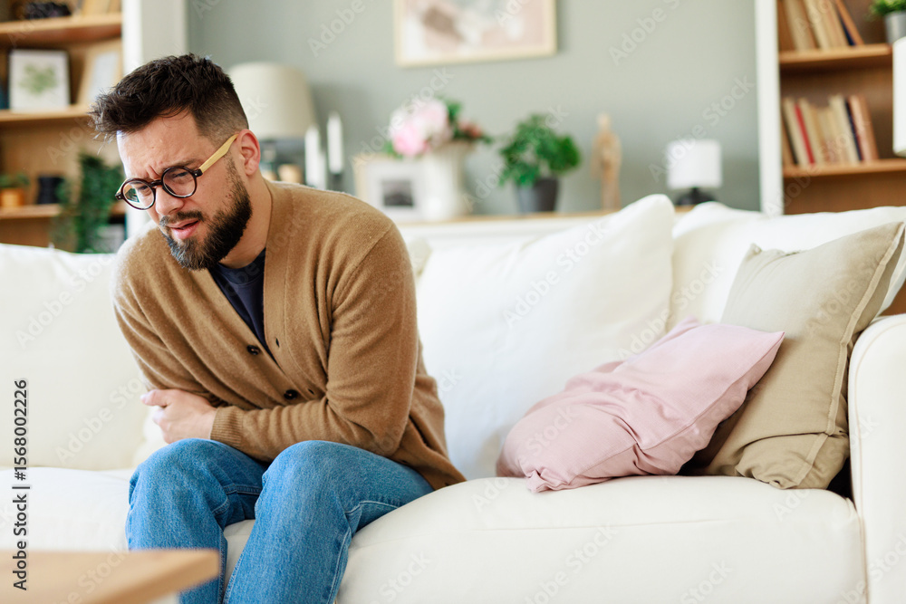 Young Caucasian Man Experiencing Pain on Sofa in Bright and Cozy Modern Living Room Young Caucasian Man Experiencing Pain on Sofa in Bright and Cozy Modern Living Room